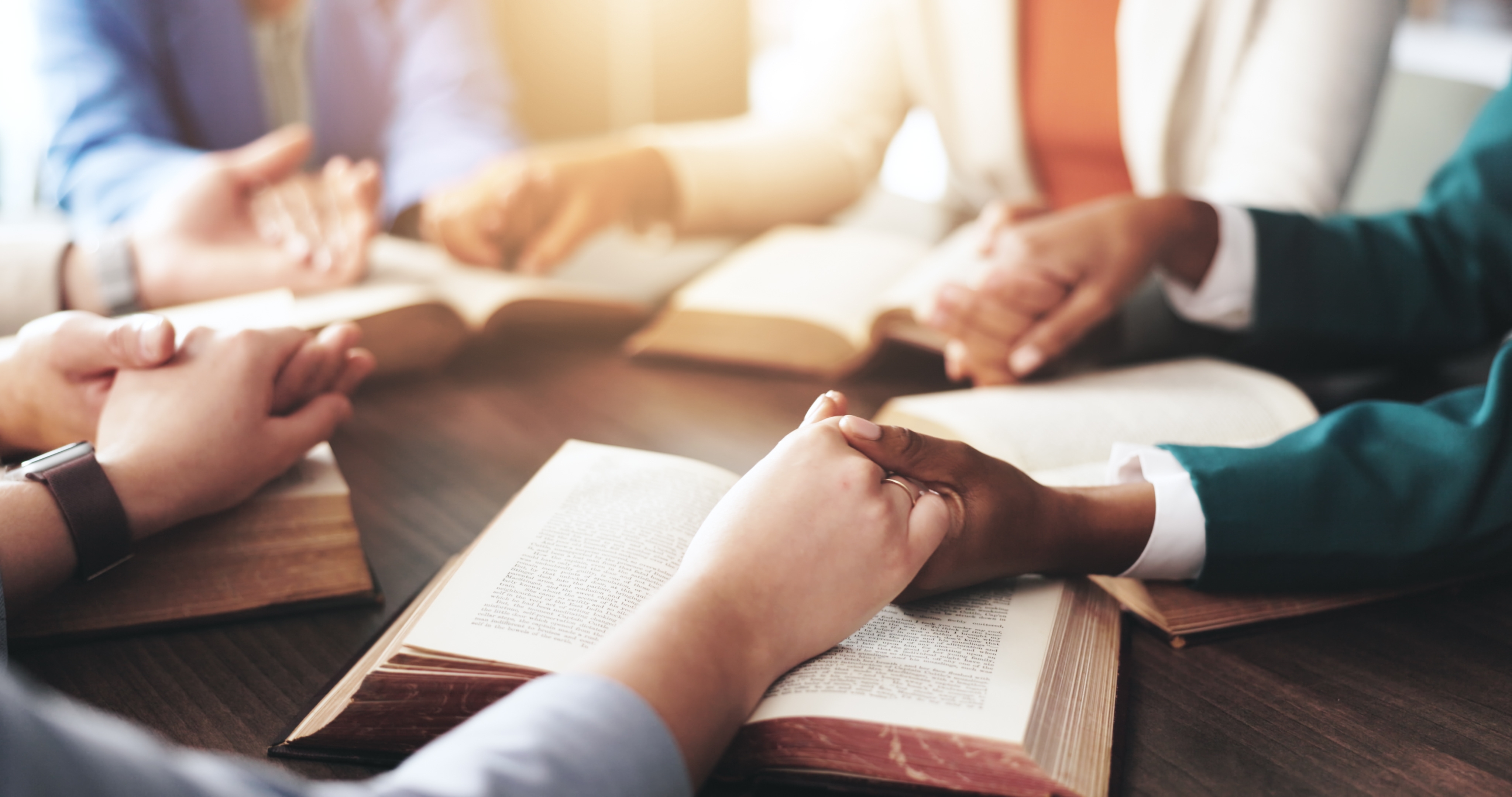 Women holding hands in prayer around open Bibles during a gathering